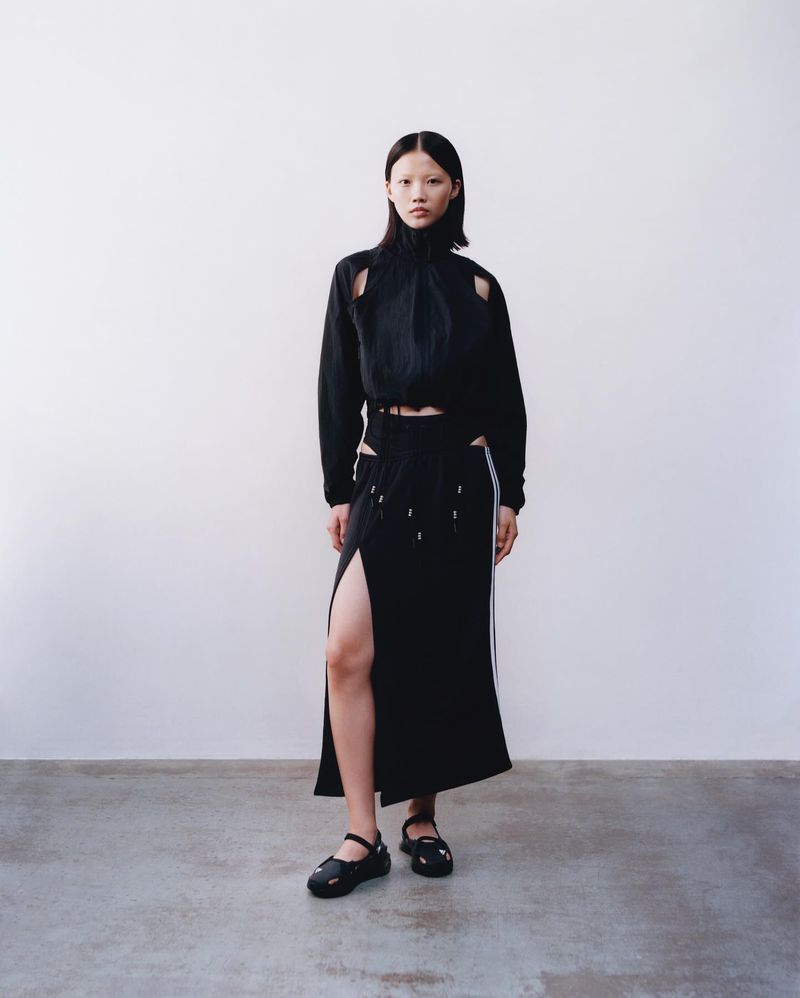 Woman posing in black cutout top and slit skirt against a white wall, showcasing modern fashion in a studio setting with neutral tones.