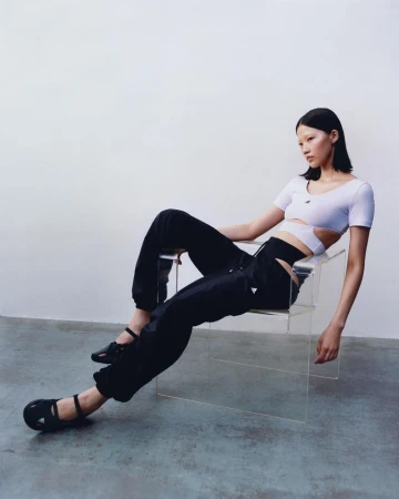 Asian woman in a cut-out white top and black pants sits in a transparent acrylic chair against a light grey studio backdrop.