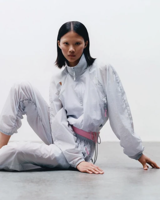 Woman with dark hair wearing a light grey tracksuit, sitting on a grey floor against a white background in a studio setting.