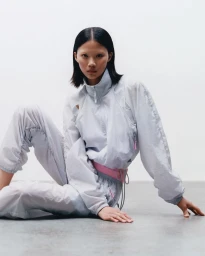 Model with dark hair wearing pale track jacket and matching trousers seated against a grey studio backdrop