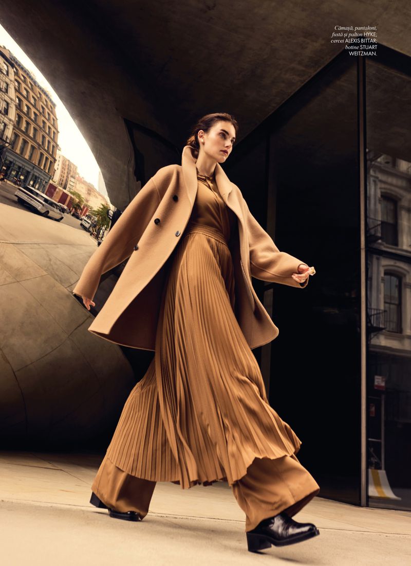 A woman in a tan coat and pleated brown dress walks on a city sidewalk. A modern building curves in the background in an urban setting.