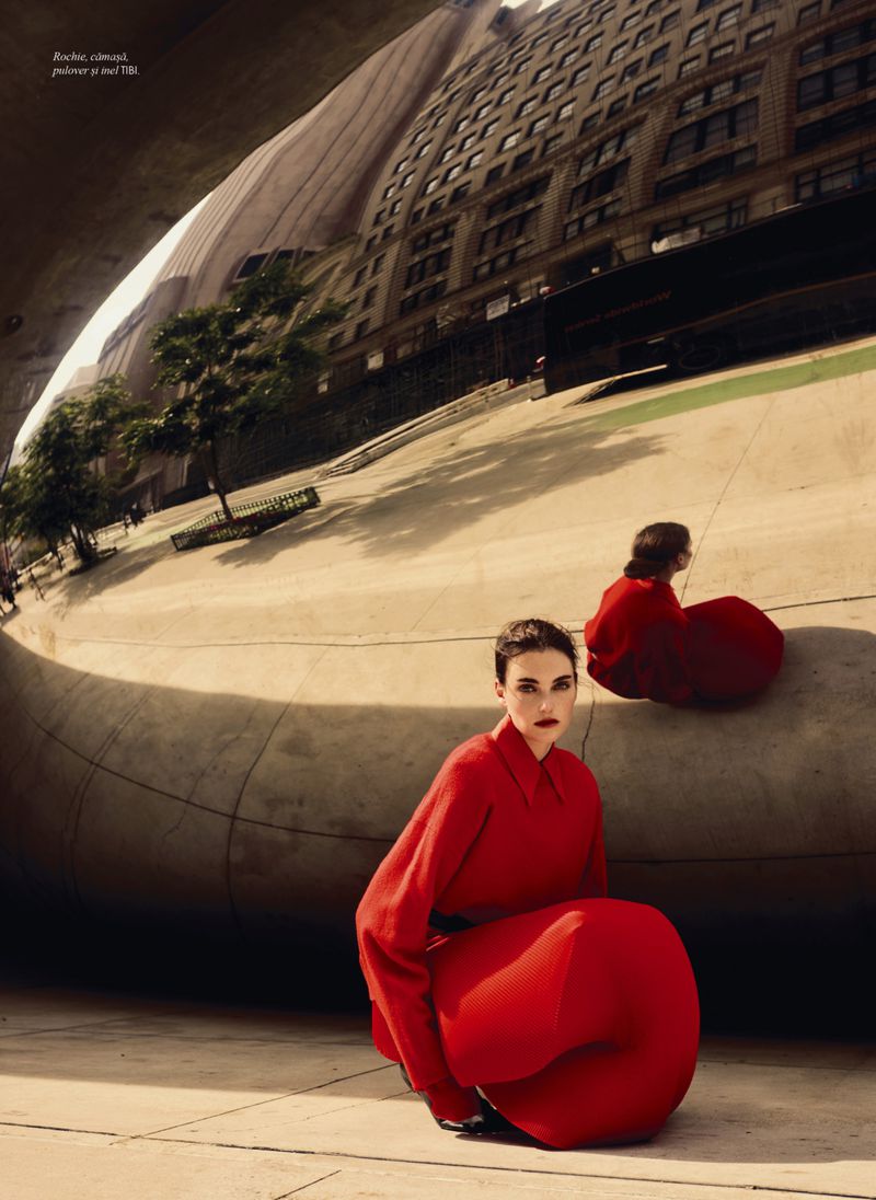 Fashion shot of a woman in a red dress kneeling in front of a mirrored sculpture reflecting city buildings and trees in an urban setting.