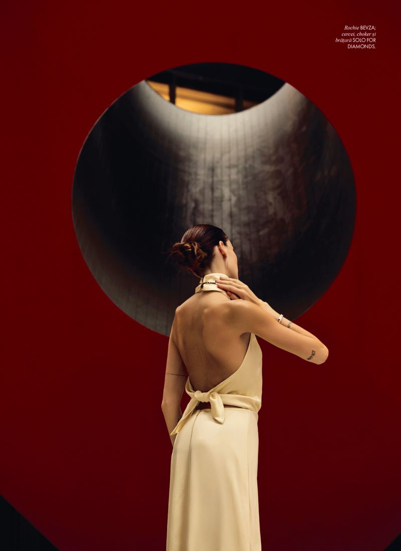 Woman in cream halter dress poses in front of a large black circle against a red background, looking upwards in an editorial fashion shot.