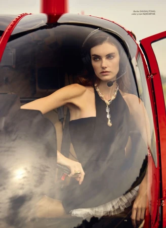 Elegant woman in a black dress sitting inside a red helicopter, wearing a headset and pearls, creating a striking fashion editorial image.