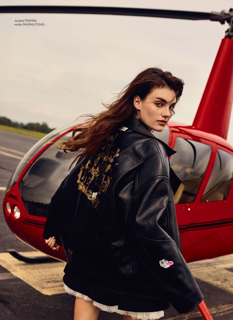 Woman in a black leather jacket stands in front of a red helicopter on an airfield, runway, or helipad on an overcast day.