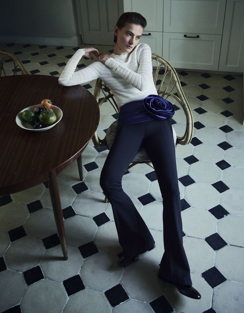 A woman in a white sweater and dark pants with a flower detail poses indoors near a dark wood table with fruit.