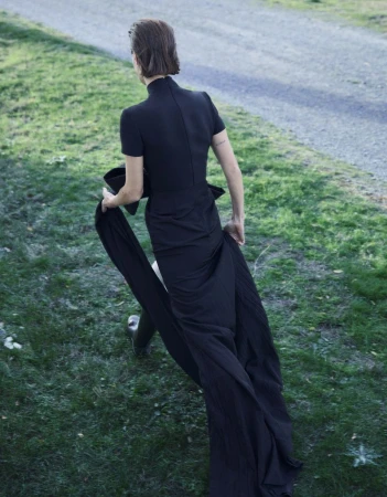 Woman in black dress walks on a grassy path. The dress has a long train. A gravel road and foliage are visible in the background.
