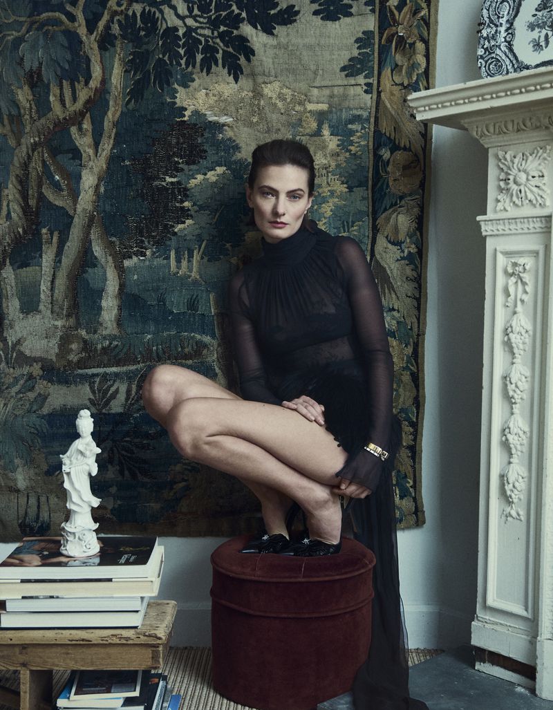 Portrait of a slender woman in a dark sheer dress, crouching on a stool in an indoor setting with a tapestry and fireplace.