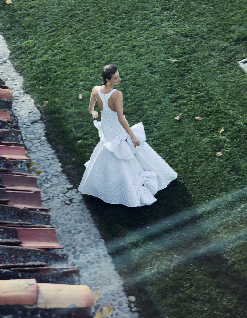 Woman in flowing white gown with oversized bow stands on stone path surrounded by green lawn and red-roofed buildings, shot from high angle.