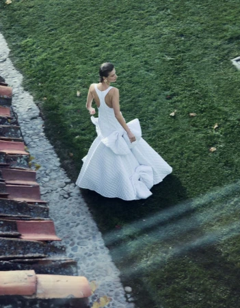 Woman in flowing white gown with oversized bow stands on stone path surrounded by green lawn and red-roofed buildings, shot from high angle.