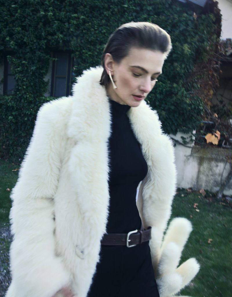 A woman with short hair is standing outdoors wearing a cream-colored fur coat, black turtleneck dress, and belt, against a green foliage backdrop.