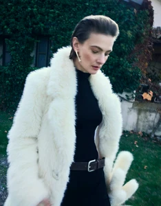Model Tanya Ruban looking downward while wearing a plush cream faux fur coat over a black turtleneck outdoors.