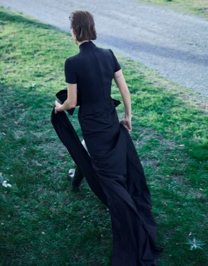 Model Tanya Ruban seen from behind walking in a long black high-neck gown across grass alongside a gravel pathway.