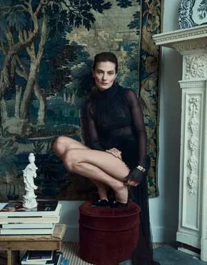 Model crouching on a velvet stool wearing a sheer black top in a room with a patterned tapestry wall hanging