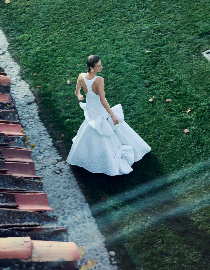 Model wearing a white structured gown walking on a lush green lawn next to a tiled rooftop edge