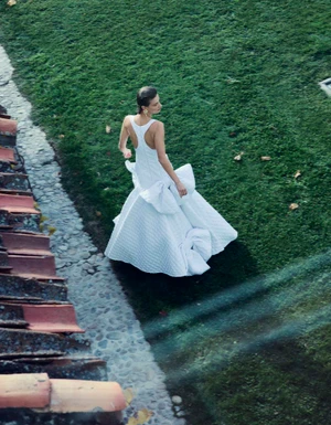 Model wearing a white structured gown walking on a lush green lawn next to a tiled rooftop edge