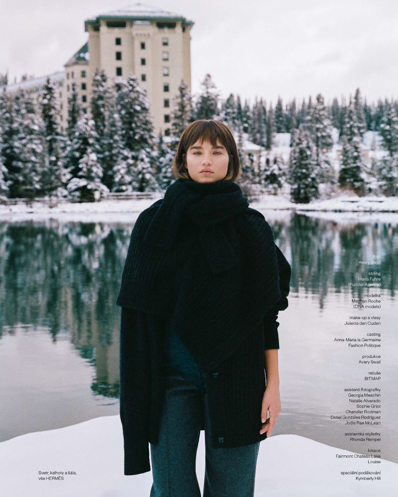 A fashionable young woman stands outdoors in winter with a snow-covered landscape, lake, and a large building in the background.