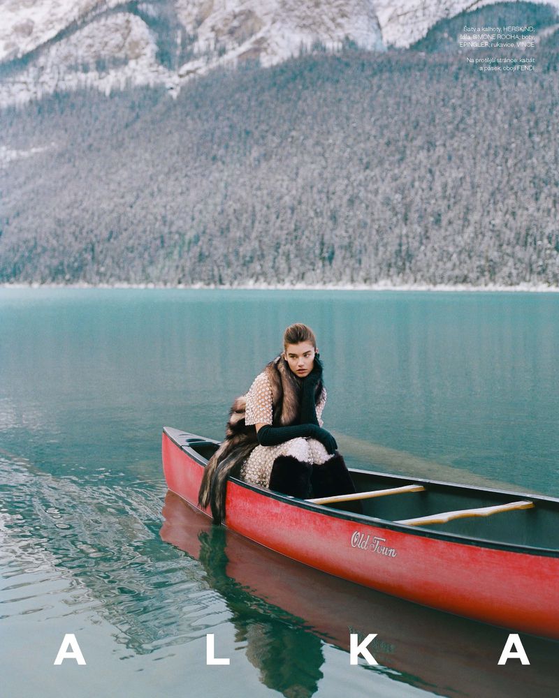 A woman sits in a red Old Town canoe on a blue glacial lake, wearing a fur coat, in front of snowy mountains and trees.