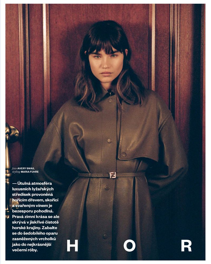 Portrait of a brunette woman wearing a brown button-up dress with a belt and Fendi buckle standing indoors in front of a dark wood door.