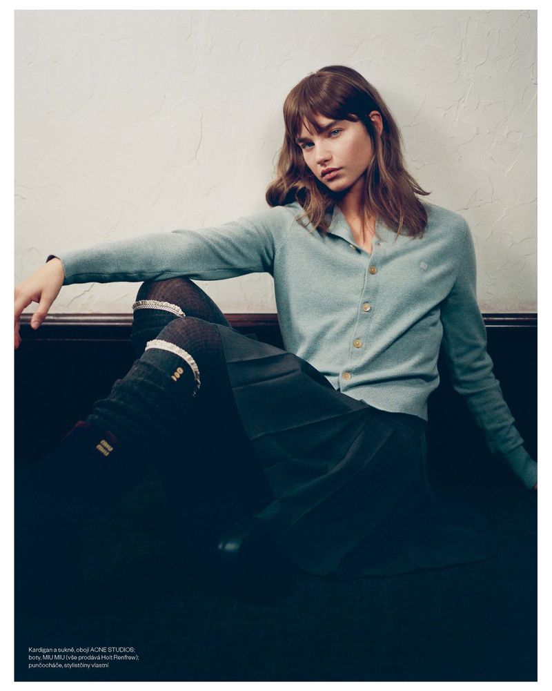 A young woman with long brown hair poses indoors wearing a light blue cardigan, dark skirt and knee-high socks in a fashion editorial.