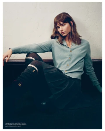 A young woman with long brown hair poses indoors wearing a light blue cardigan, dark skirt and knee-high socks in a fashion editorial.