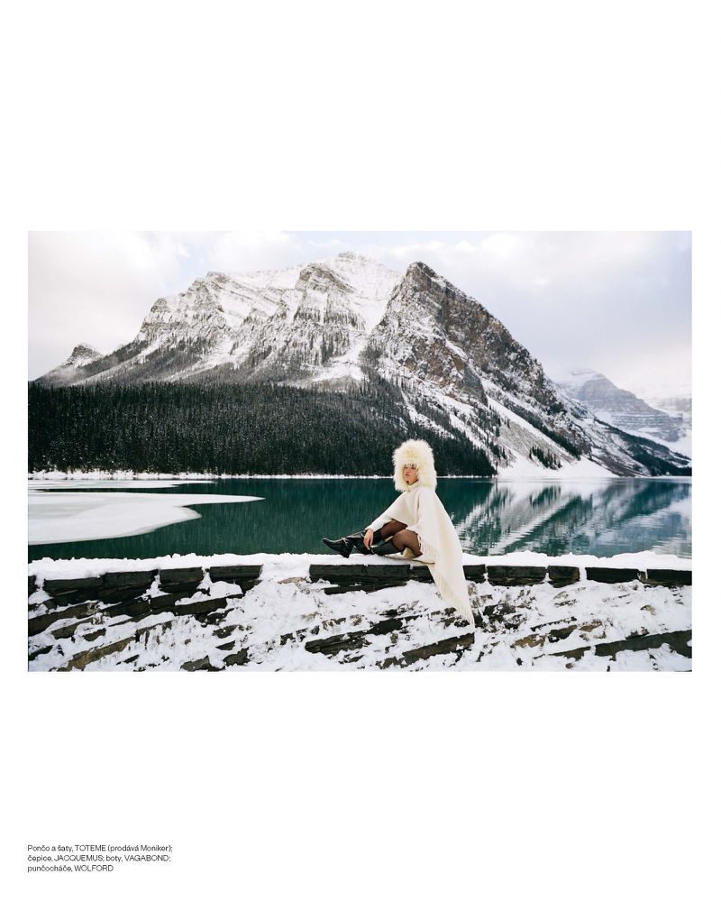 A fair-skinned adult female model in white fur hat and cape poses on a snow wall with a large mountain and turquoise lake background.