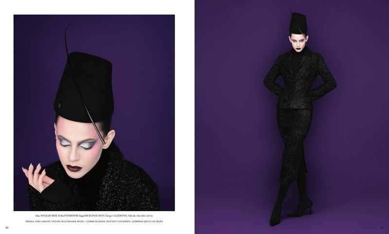 Fashion editorial shot featuring a woman in a black suit and matching hat, against a bold purple backdrop, highlighting dark makeup and styling.
