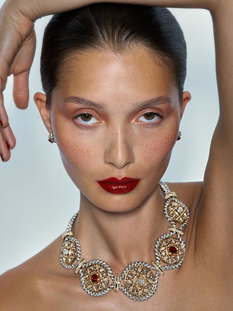 Beauty portrait of model Yada Villaret wearing bold red lipstick and an ornate gold statement necklace against a pale background.