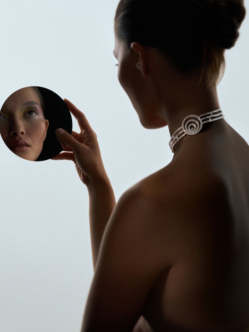 Adult woman poses with a round mirror, showing her reflection, wearing a necklace with a circular design, against a light gradient backdrop.