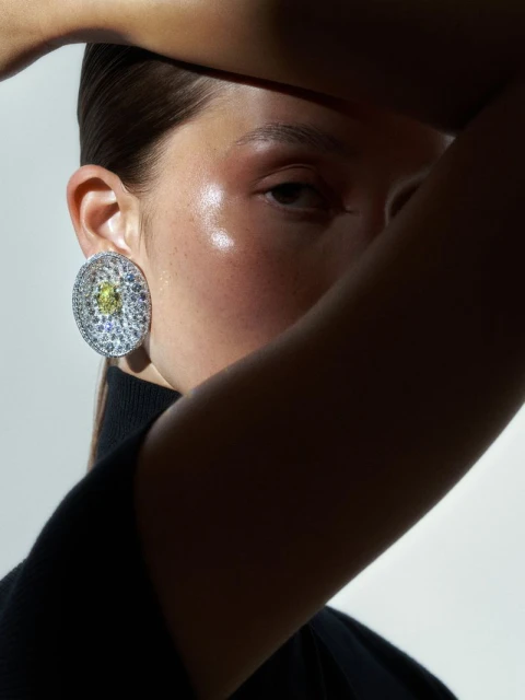 Woman wearing a silver earring with a large gemstone and shielding her face from the sun, casting shadows in a bright setting.