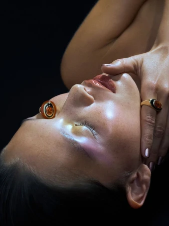 Close-up portrait of a model with closed eyes wearing two statement cocktail rings against a dark background.