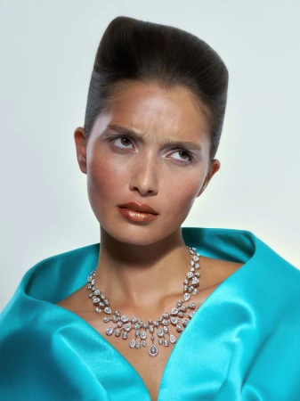 Woman with high hairstyle and blue silk shawl poses with a diamond necklace against a neutral background in a fashion portrait.