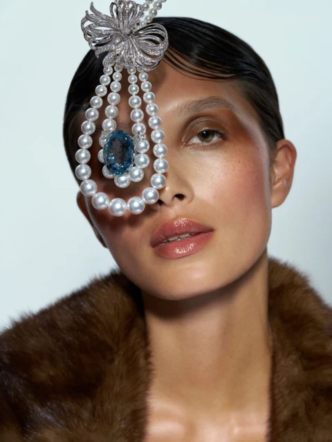 Close-up of a woman with a pearl and blue gem headpiece, wearing a fur coat, glossy lips, and wet-look hair against a light background.