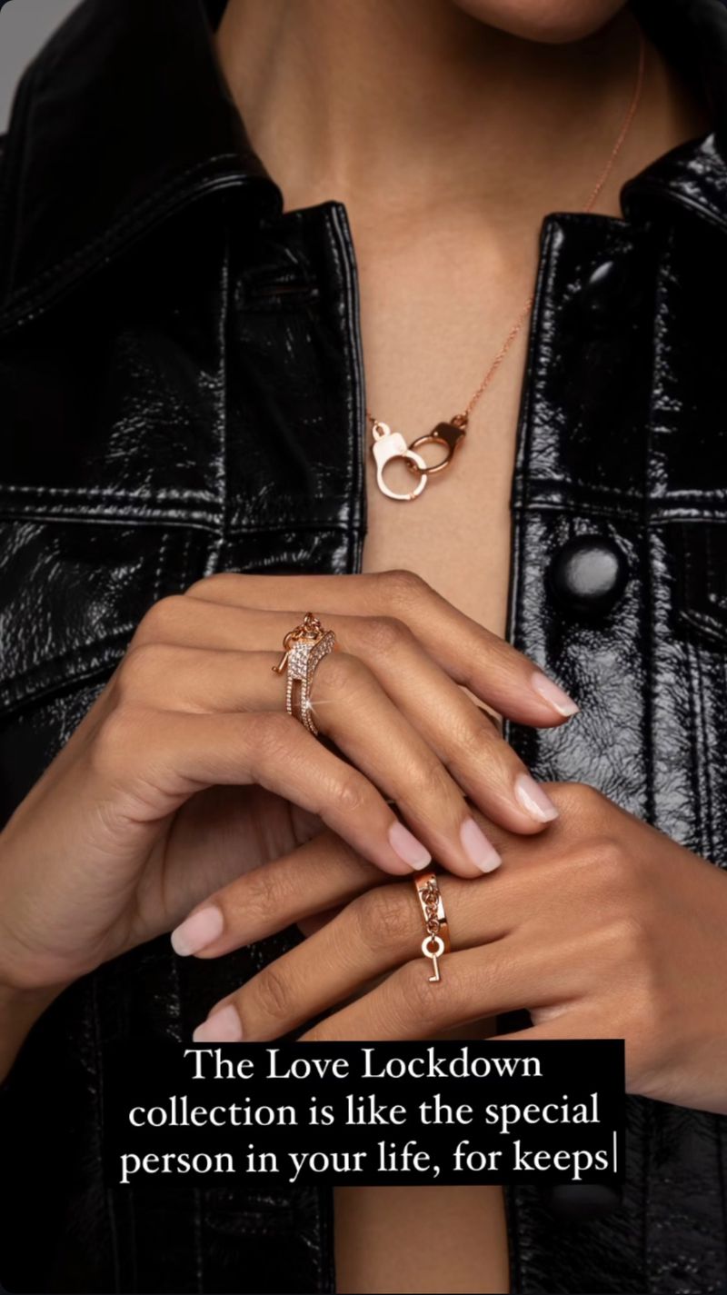 Close-up of a woman's hands displaying gold rings, with a gold handcuff necklace. The model wears a shiny black jacket.
