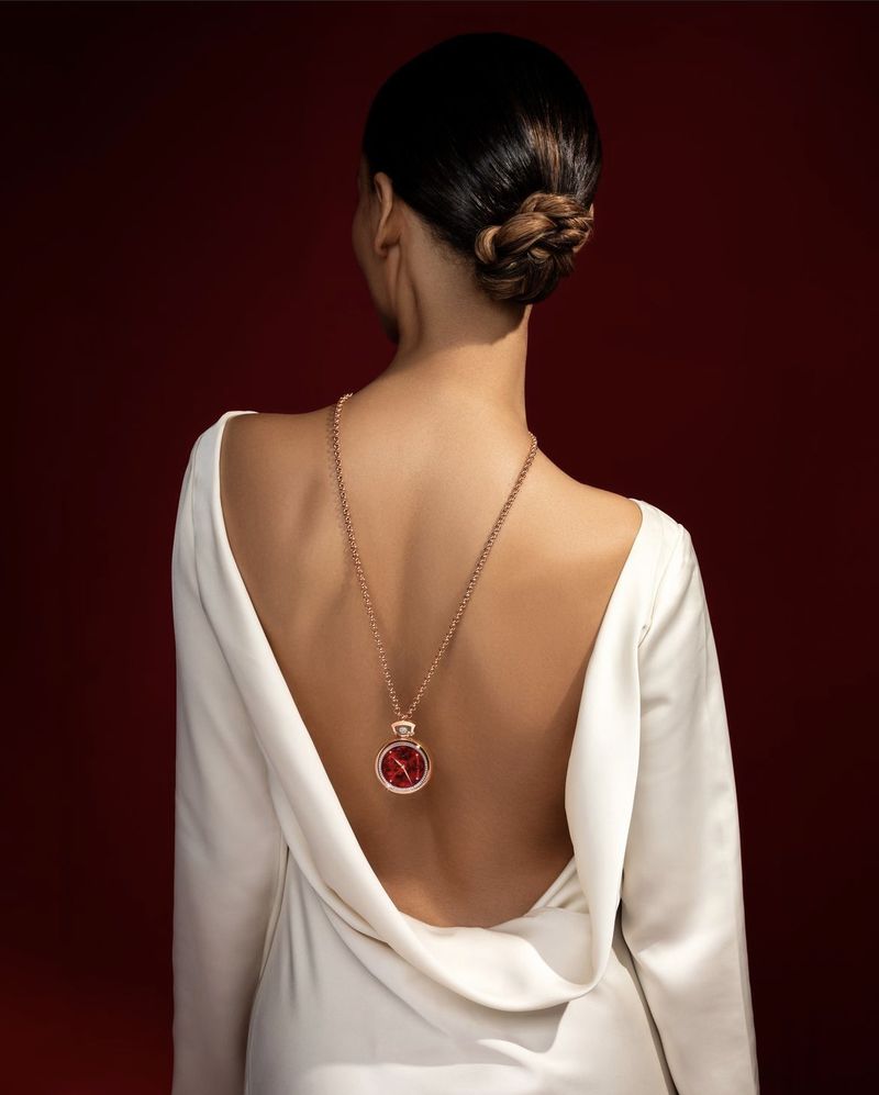 An adult female stands with her back to the camera, wearing a white dress and a long gold necklace with a red pendant.