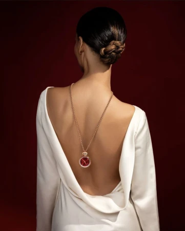 An adult female stands with her back to the camera, wearing a white dress and a long gold necklace with a red pendant.