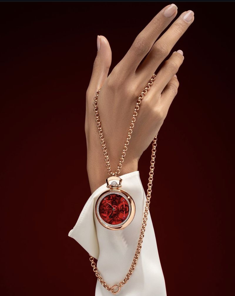 Woman's hand holds a round pendant watch with a gold chain, on a white fabric and a dark red backdrop in a beauty fashion shot.