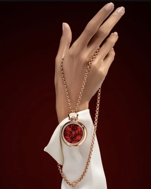 Woman's hand holds a round pendant watch with a gold chain, on a white fabric and a dark red backdrop in a beauty fashion shot.