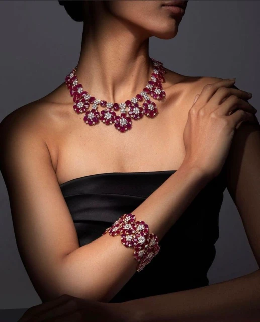 Woman wearing a floral ruby and diamond necklace with matching bracelet. The jewelry is set against a gradient dark gray background.