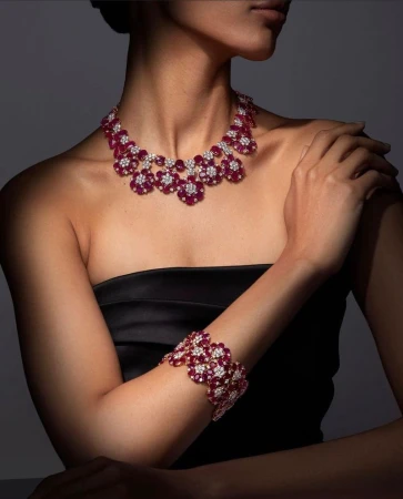 Woman wearing a floral ruby and diamond necklace with matching bracelet. The jewelry is set against a gradient dark gray background.