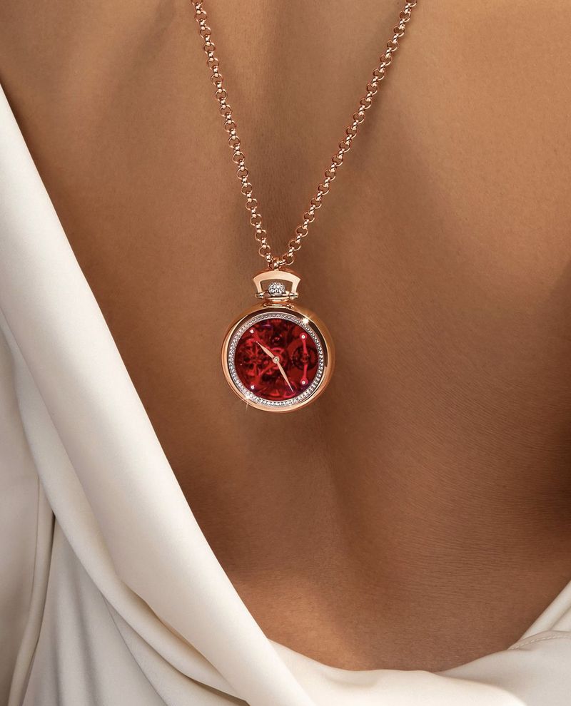 Close up of a rose gold necklace with a red watch pendant worn against a model's back in a white silk dress