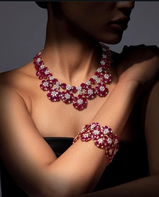 Close-up studio shot of a woman displaying a red and white floral-patterned ruby and diamond necklace and bracelet jewelry set.