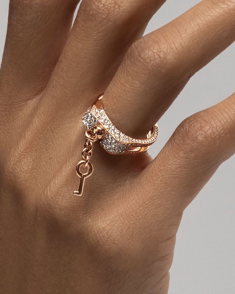 Close-up of a woman's hand wearing a rose gold ring with diamonds and a dangling key charm against a soft neutral background.