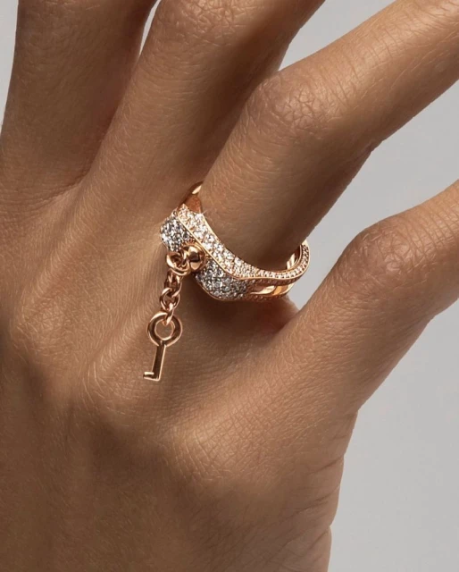 Close-up of a woman's hand wearing a rose gold ring with diamonds and a dangling key charm against a soft neutral background.