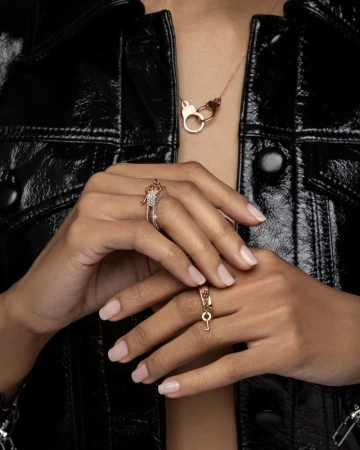 Close-up of hands displaying gold diamond rings and matching handcuffs pendant necklace against a glossy black jacket