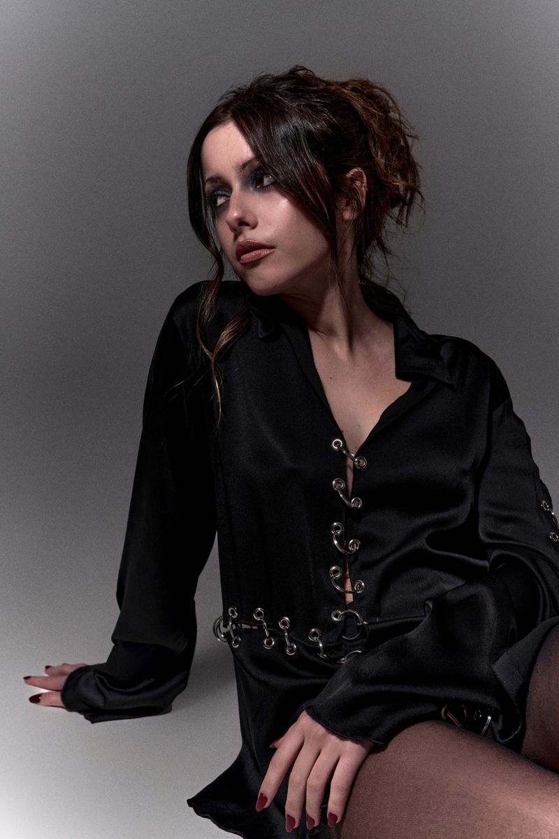 Portrait of a young woman in a black satin shirt with metal ring details, sitting indoors with a soft studio lighting, looking away.