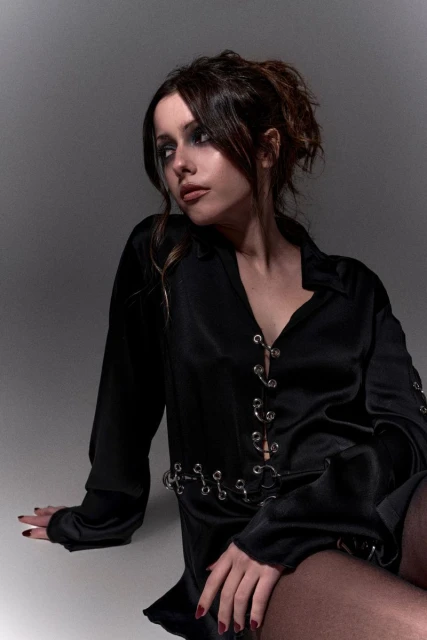 Portrait of a young woman in a black satin shirt with metal ring details, sitting indoors with a soft studio lighting, looking away.