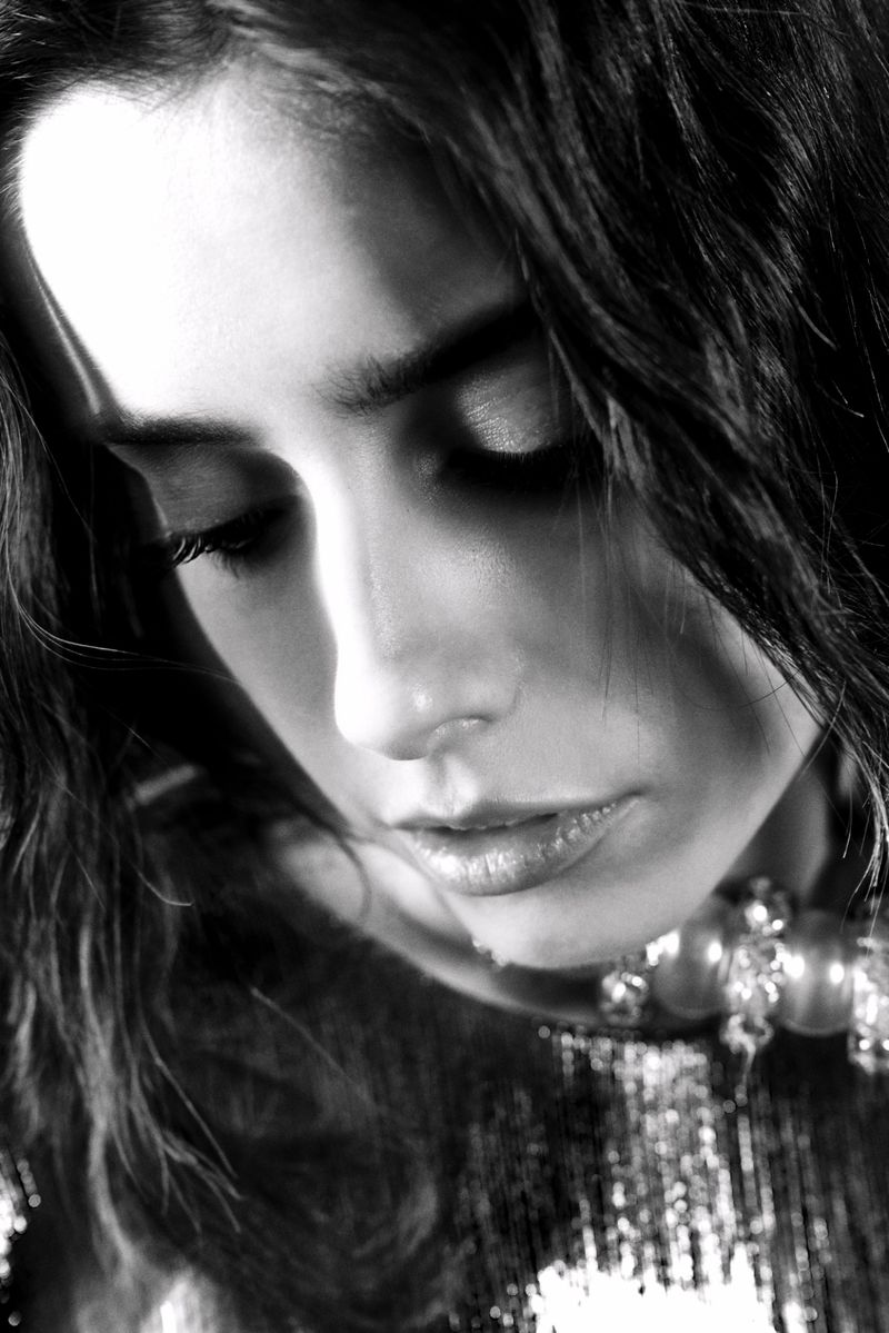 Monochrome portrait of a woman with wavy dark hair gazing downward, wearing a beaded necklace and shimmering top in soft light.