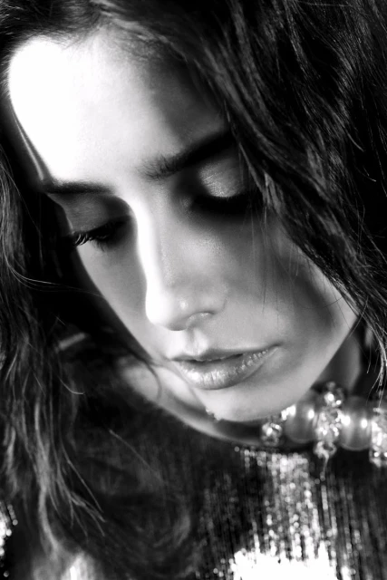 Monochrome portrait of a woman with wavy dark hair gazing downward, wearing a beaded necklace and shimmering top in soft light.