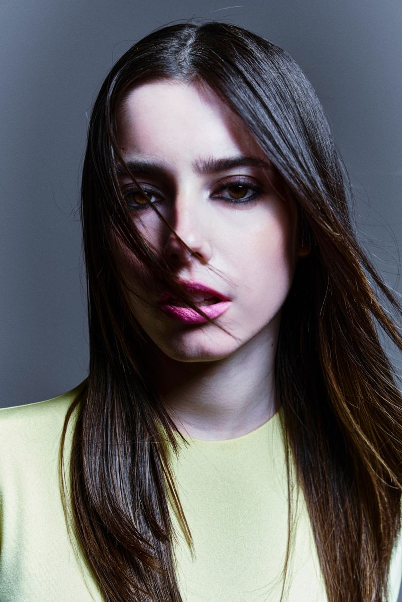 Close-up portrait of a young woman with long dark hair and strands across her face, wearing a yellow top against a grey studio background